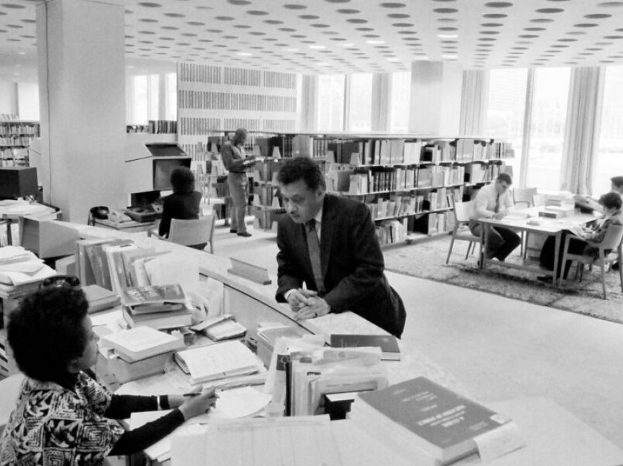 Biblioteksmiljø i sort-hvid: en person taler med en bibliotekar ved en skranke, mens andre læser ved borde.